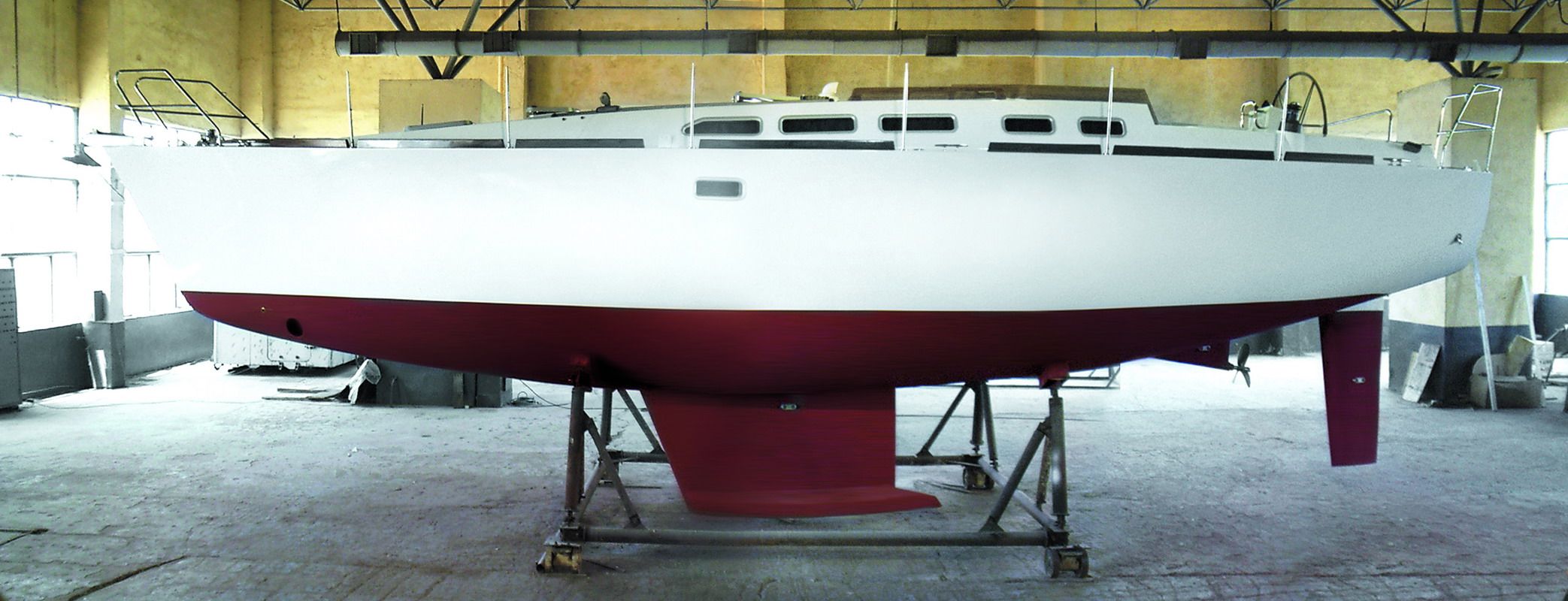 The steel round-bilge hull of the Madeira 44 in profile in the build hall — white topsides over red antifouling, fin keel and skeg-hung rudder.