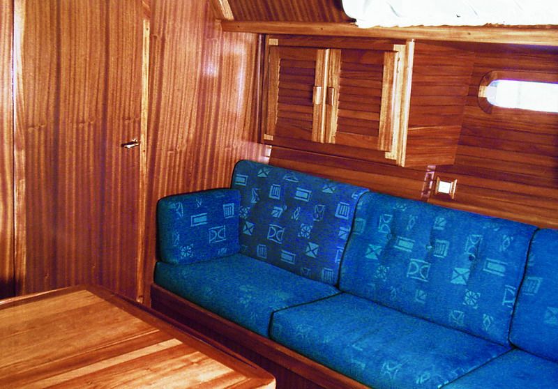 A saloon settee berth in blue upholstery below a hull portlight, in solid mahogany.